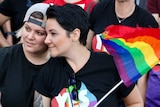 One woman with a backwards cap rests her head on another woman, who is holding a rainbow flag.