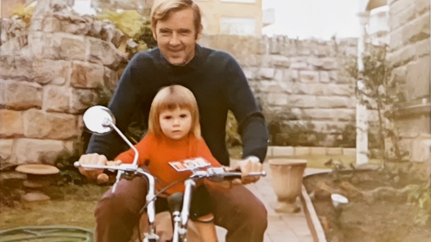 a little girl in a red jumper is sitting on a motorbike with an adult man weraing a blue jumper