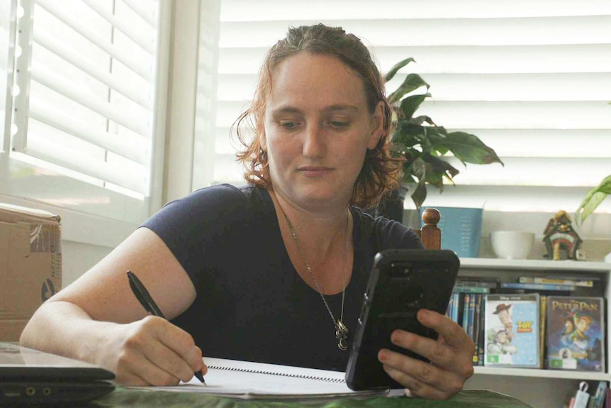 Bianca Hackett sitting at a desk next to shuttered windows holding a mobile and writing in a book
