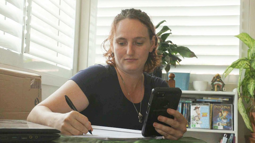 Bianca Hackett sitting at a desk next to shuttered windows holding a mobile and writing in a book