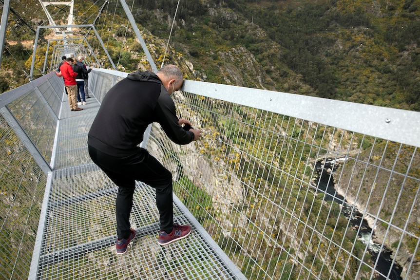 Një burrë bën një foto të një lumi nga një urë e tejdukshme metalike.