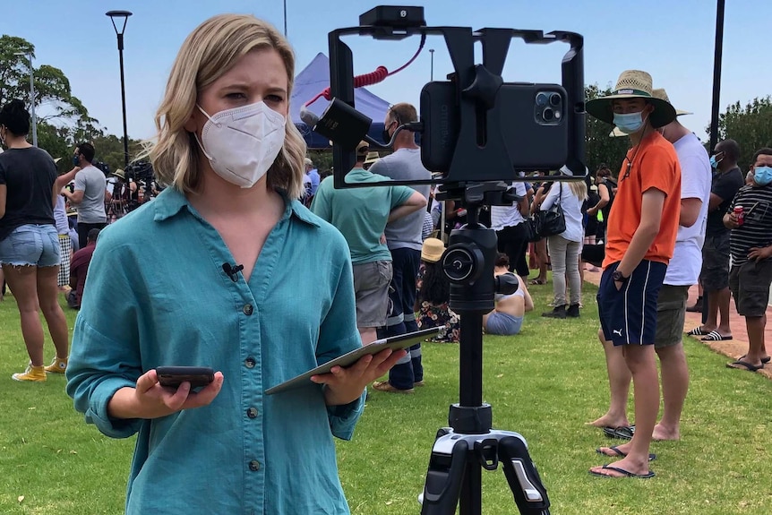 Woman wearing a mask, holding ipad talking to smart phone on a tripod.