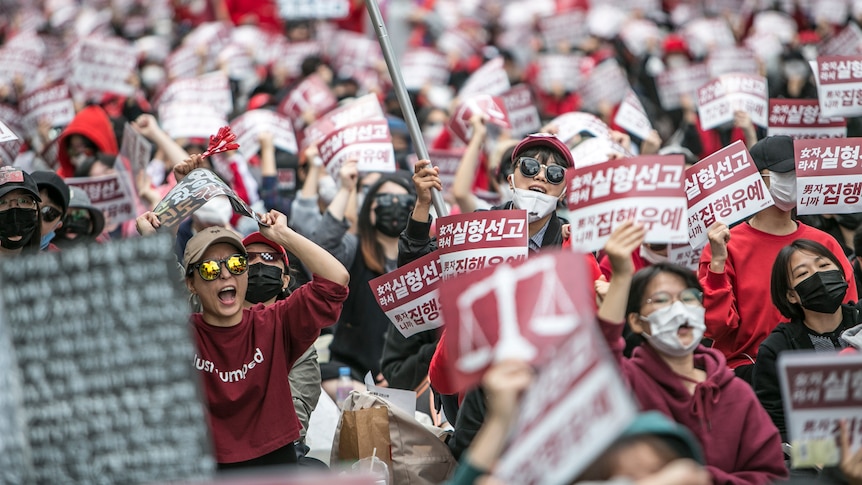 A large crowd of women wearing red hold posters and placards with Korean writing. Some are shouting.