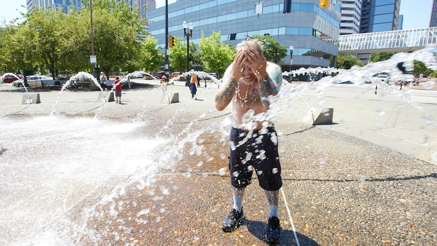 A man stands shirtless under a water fountain splashing water onto his face.