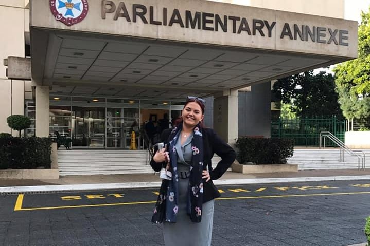 A young woman in a smart grey dress and black jacket stands outside a building with 'parliamentary annexe' on it