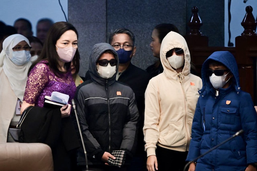 A woman wearing purple lace dress stands with three teens in hoodies, dark glasses and face masks