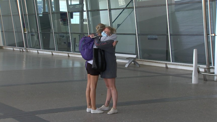 A woman wearing a mask hugs a woman carrying a large purple backpack