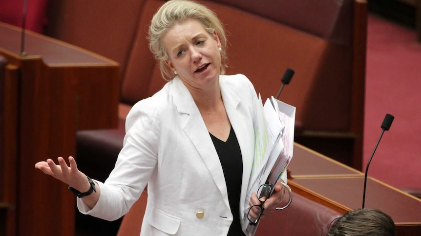 Bridget McKenzie looks across the room and holds her arms in a shrug while also carrying binders