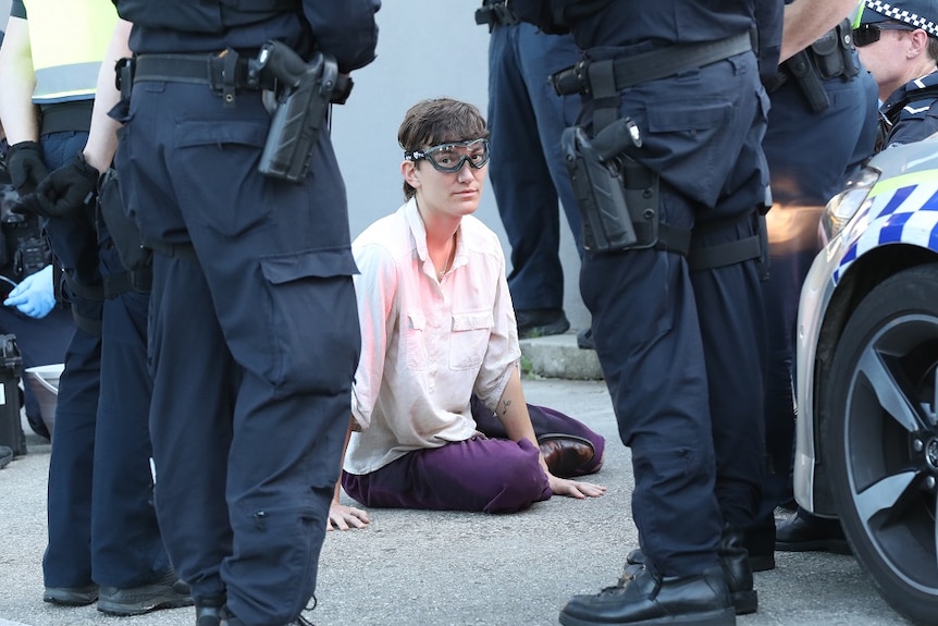 A woman wearing safety goggles sits in the middle of a driveway surrounded by police officers.