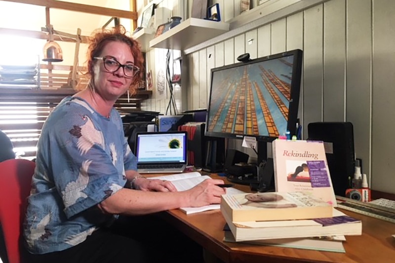 Narelle Dickinson sits at a computer at a desk with books around her in her office.