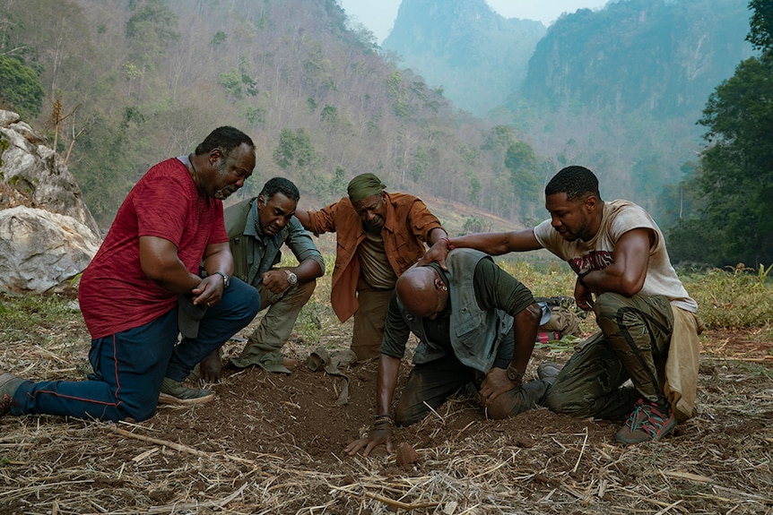 On hazy day in mountainous jungle, four men in camping attire kneel on one kneel around a distraught man kneeling in dirt ditch.