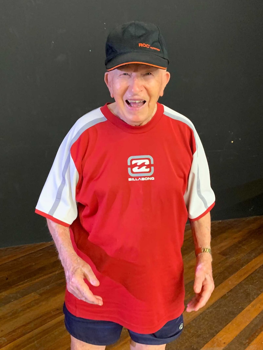 An old man in a black cap and red and white shirt smiles at the camera.