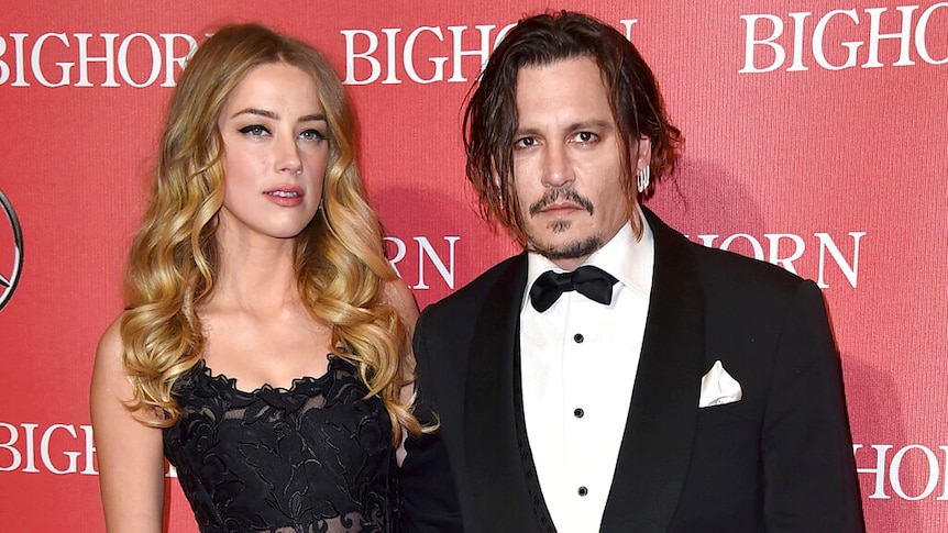 Amber Heard and Johnny Depp pose into front of a logo wall on a red carpet event, both in formal wear.