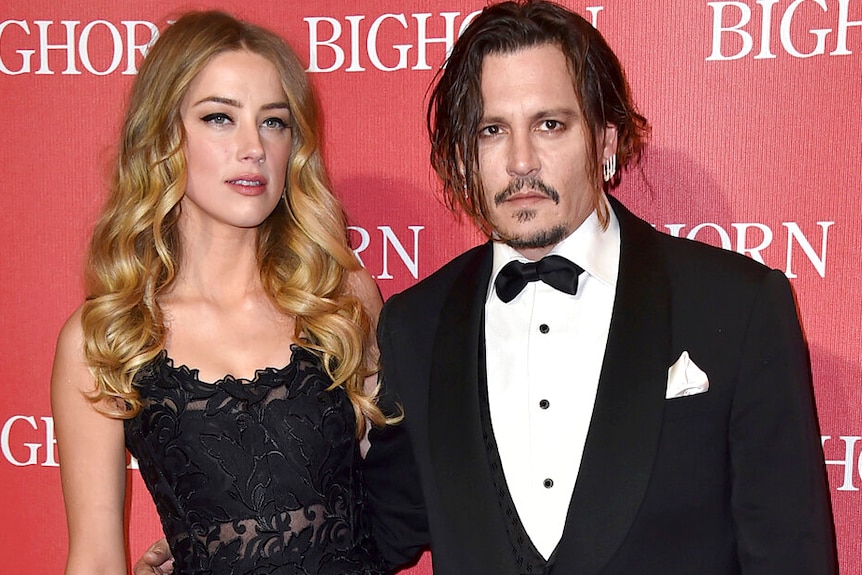 Amber Heard and Johnny Depp pose into front of a logo wall on a red carpet event, both in formal wear.