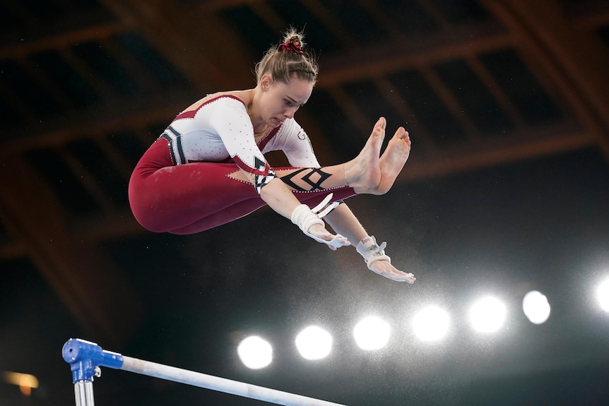 Sarah Voss mid-air reaching for her toes performing on the uneven bars.