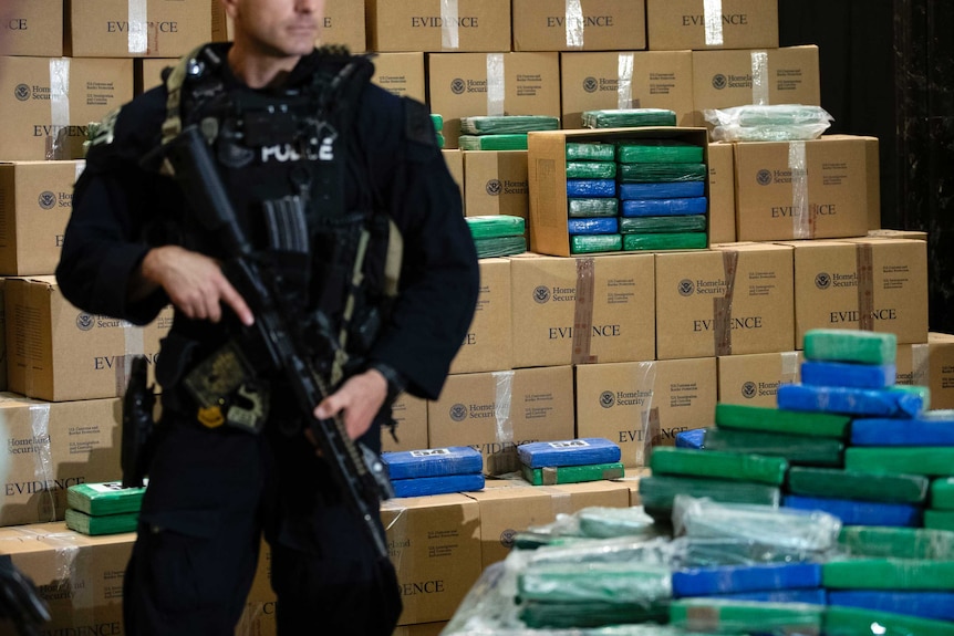 A police officer with an automatic rifle stands guard in front of boxes of cocaine.