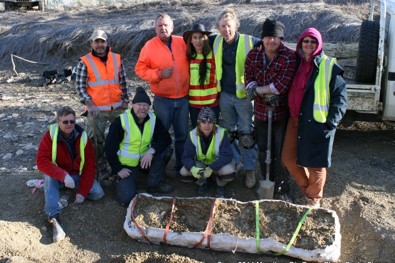 A group of people wearing hi-vis vests and causal attire stand at a dig site behind a fossil specimen on the ground