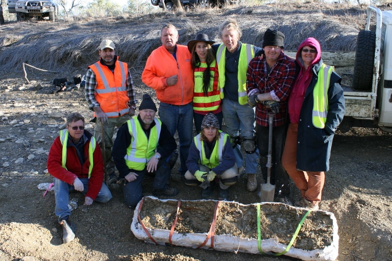 A group of people wearing hi-vis vests and causal attire stand at a dig site behind a fossil specimen on the ground