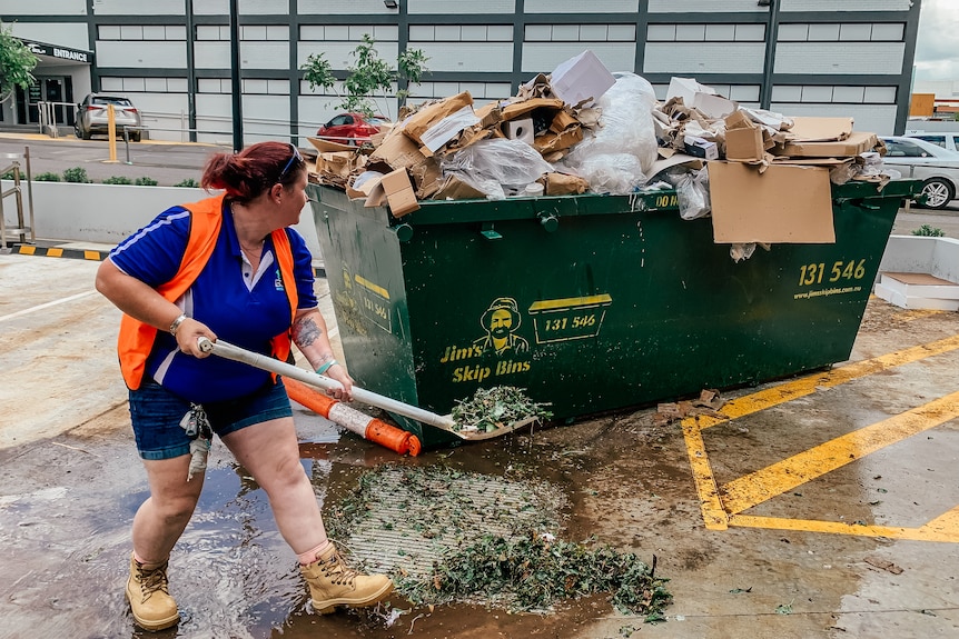 a woman shovels leaves into a very full rubbish skip