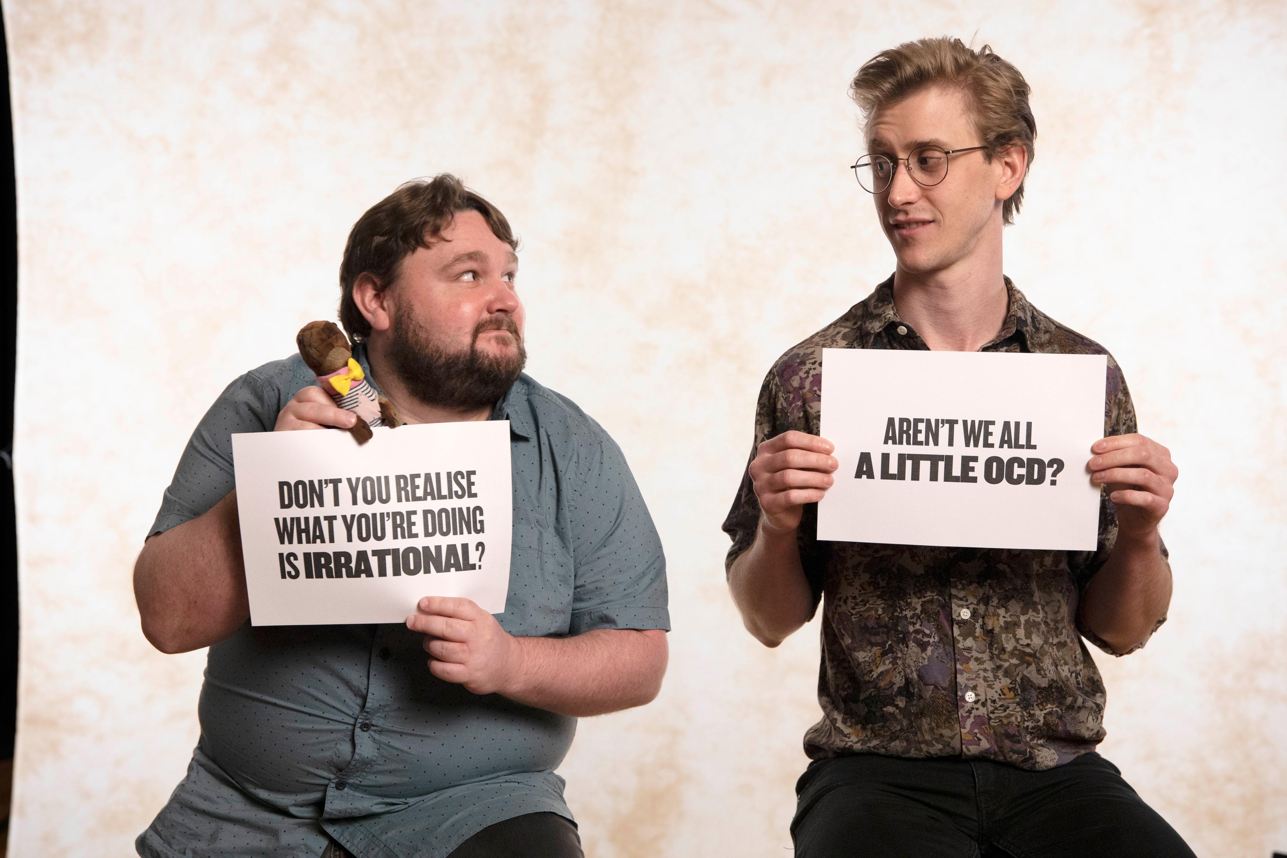 Two men sit on stools holding signs one says "aren't we all a little OCD?"