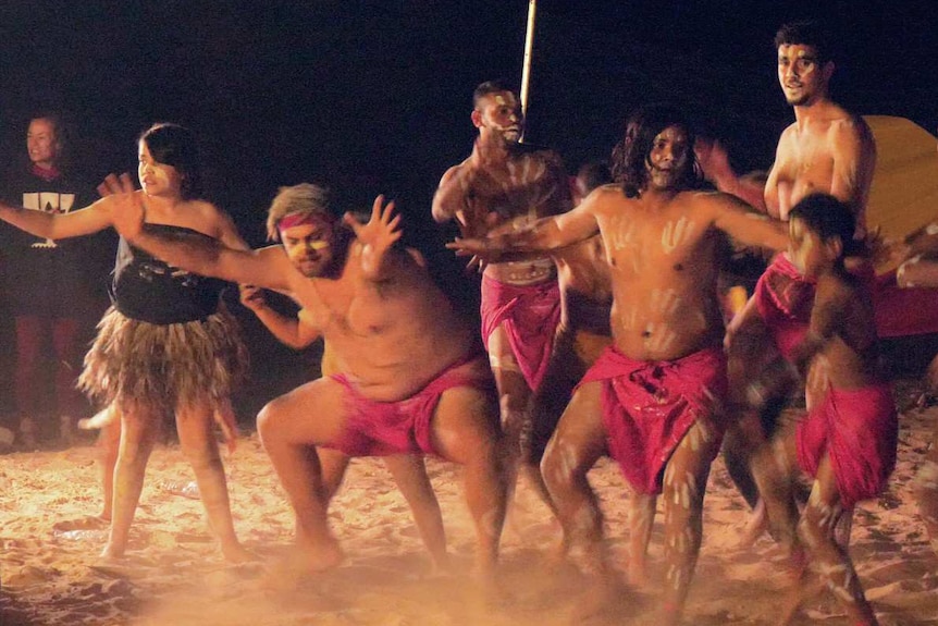 A group of Indigenous dancers in white body paint, red loin cloth or emu feathers in motion at night on a red dirt dance ground.