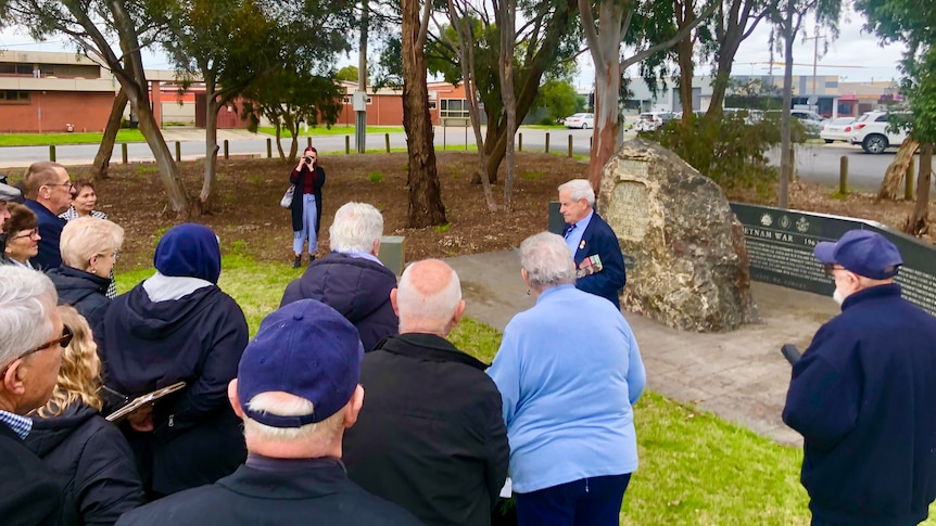 A group of people to the right listen to a man wearing medals make a speech near a grey stone war memorial on a cloudy day