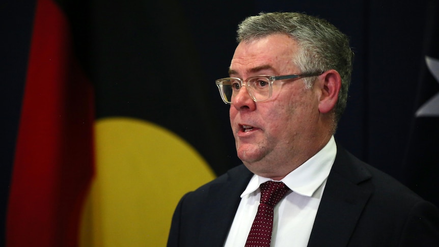 A caucasian man in a dark suit and red tie faces to the left while speaking. Behind him is an Aboriginal flag. 