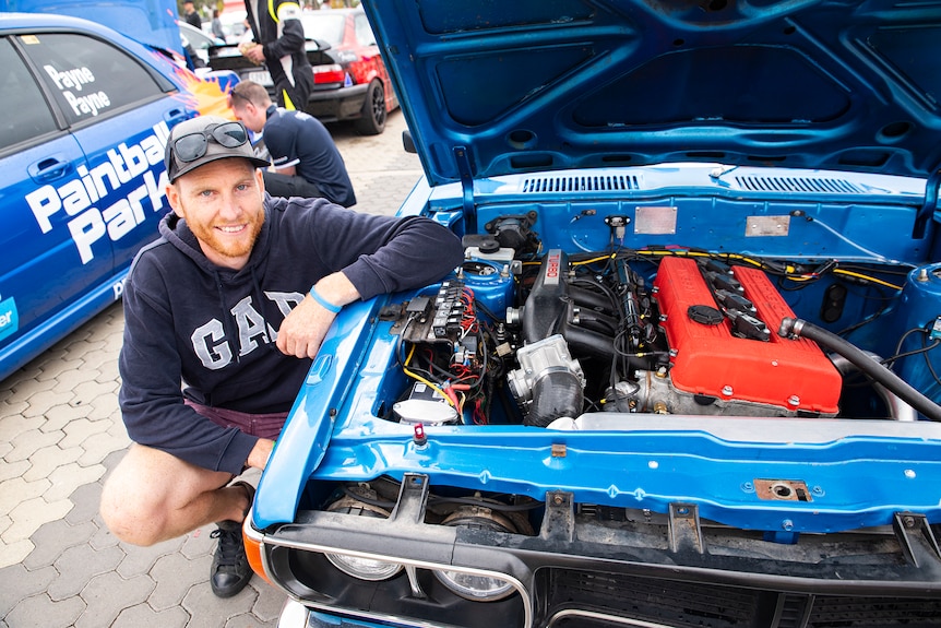 A man crouches by the open bonnet of his blue car