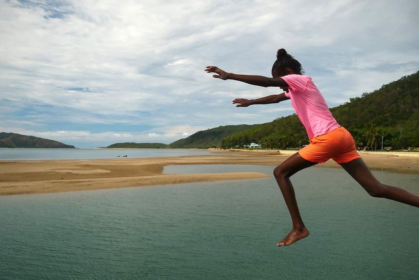 A young Aboriginal girl wearing bright swimwear jumps from the jetty on Palm Island.