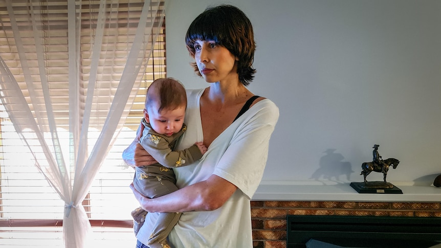 A serious young woman with short hair stands holding her a baby in a lounge room. Wears white t-shirt, looks away from camera.