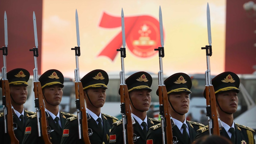 Chinese soldiers holding bayonets stand in a row in front of a screen saying 70.