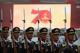 Chinese soldiers holding bayonets stand in a row in front of a screen saying 70.