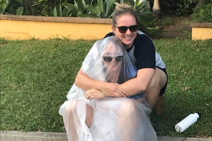 A woman hugs another woman who sits in the gutter, covered in a sheet of plastic.