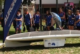 A sleek solar car on the grass outside a school surrounded by kids. Big teardrop flag banners.
