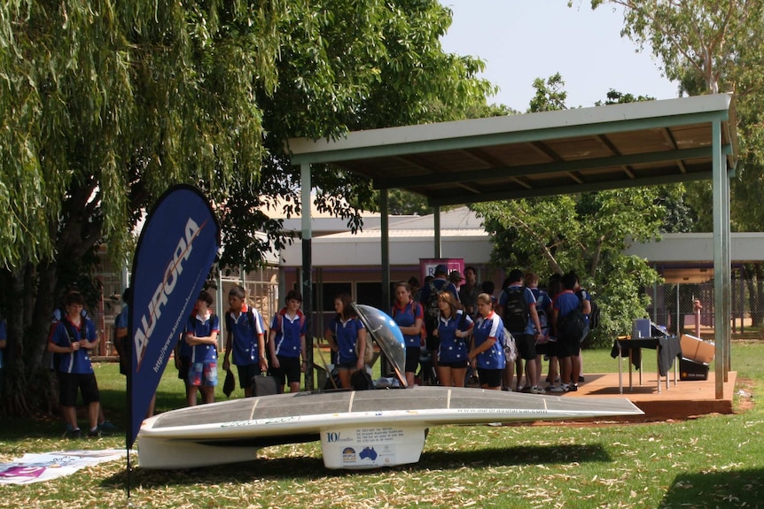 A sleek solar car on the grass outside a school surrounded by kids. Big teardrop flag banners.