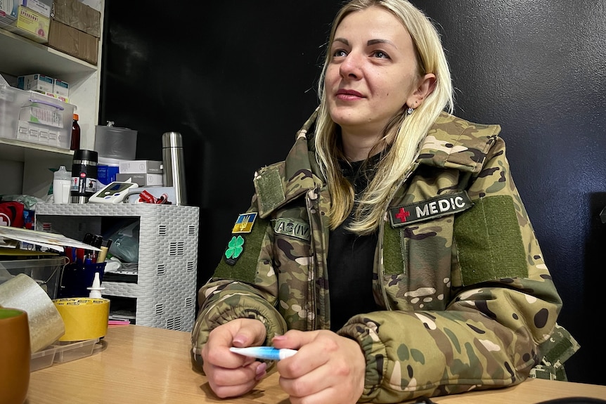 A woman with blonde hair and wearing army fatigues sits at a desk looking off camera.
