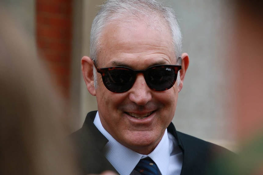 A head and shoulders shot of a smiling man with grey hair wearing dark sunglasses, a coat and shirt and tie.