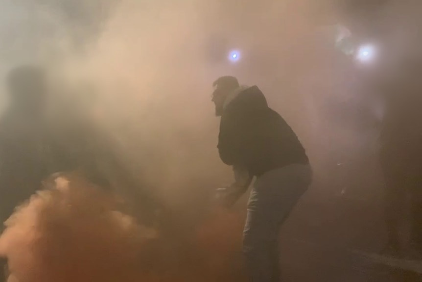 a man wearing a hooded jumper can be seen through the mists of orange flare smoke at the Sydney Opera House