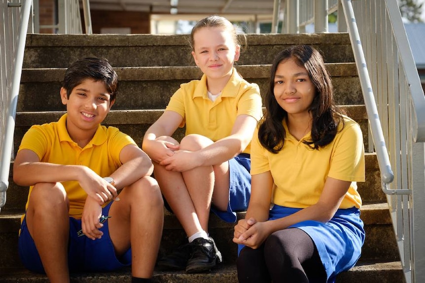 Three primary school aged children in gold and blue uniforms sitting on a concrete staircase
