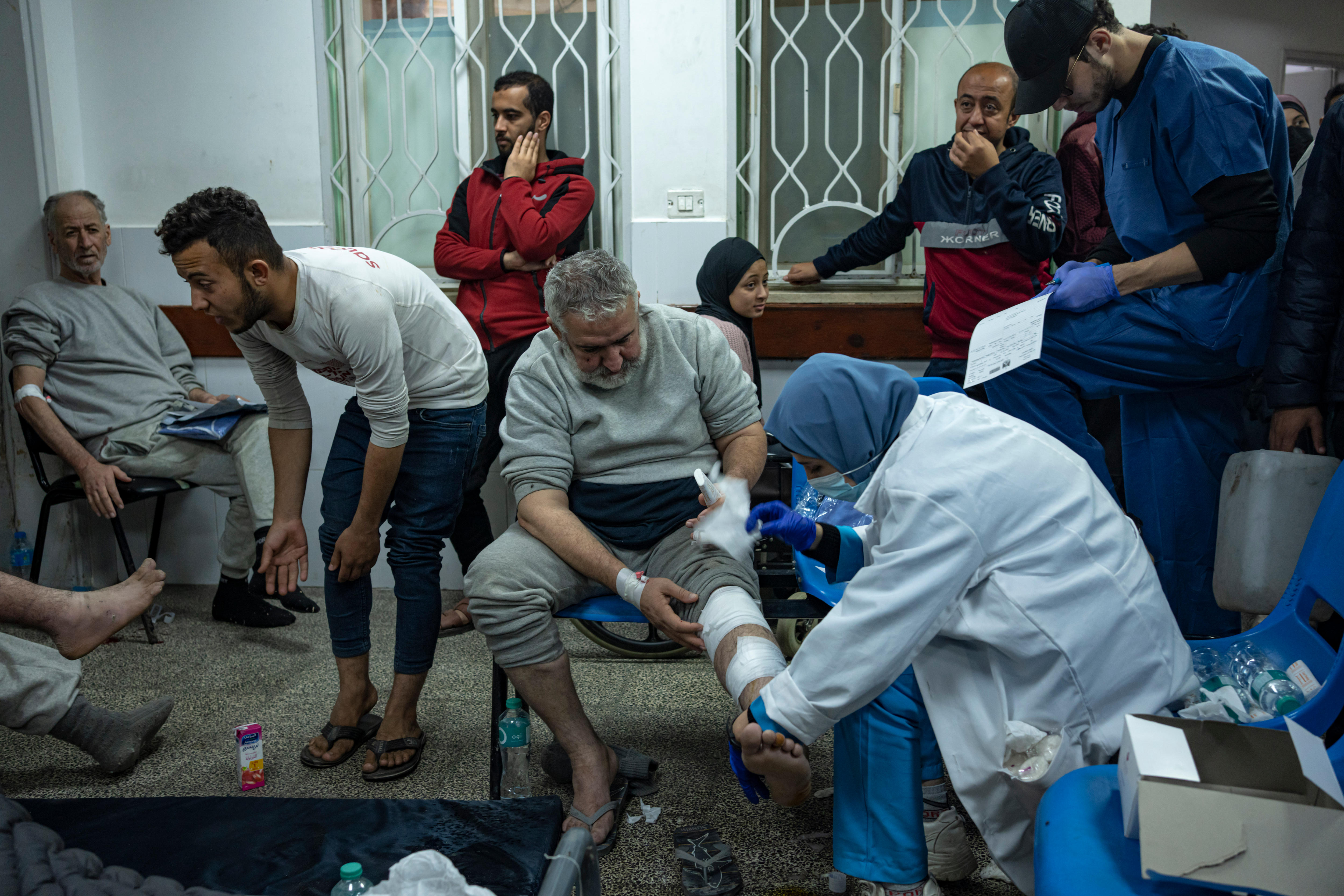 Man sits on chair while doctor wraps his leg in a bandage. Other men sit and stand in the background. 
