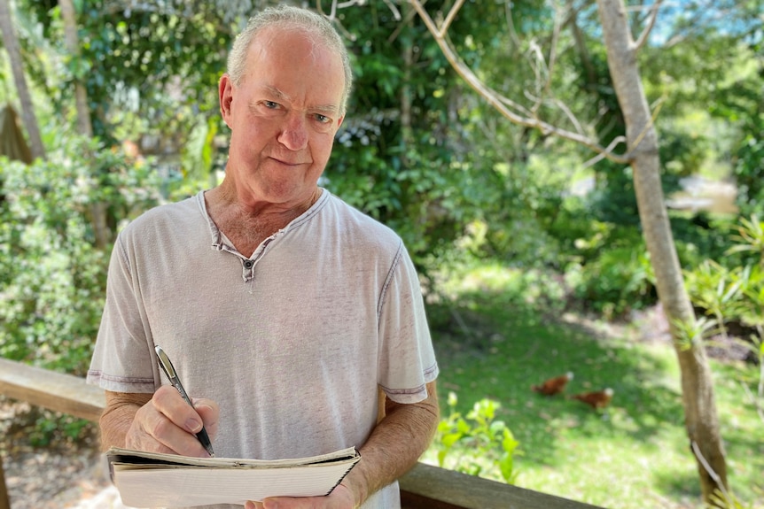 Man in his 70s with a pen and paper on a deck, with greenery and chooks in background