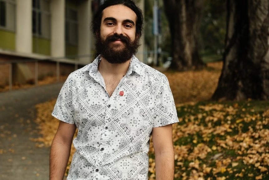 A man with dark hair and beard stands in front of a tree