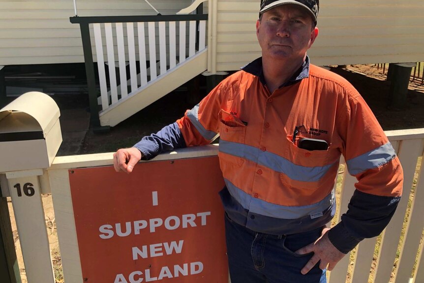 A man in high-vis stands outside a house with an "I support New Acland Mine" sign on the gate.