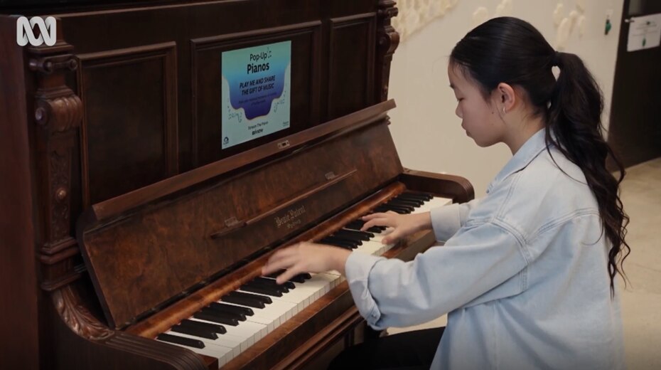 Pianist Alice, sitting on a piano stool playing a song on the brown upright piano, that has an ABC Pop Up Piano stick on it.