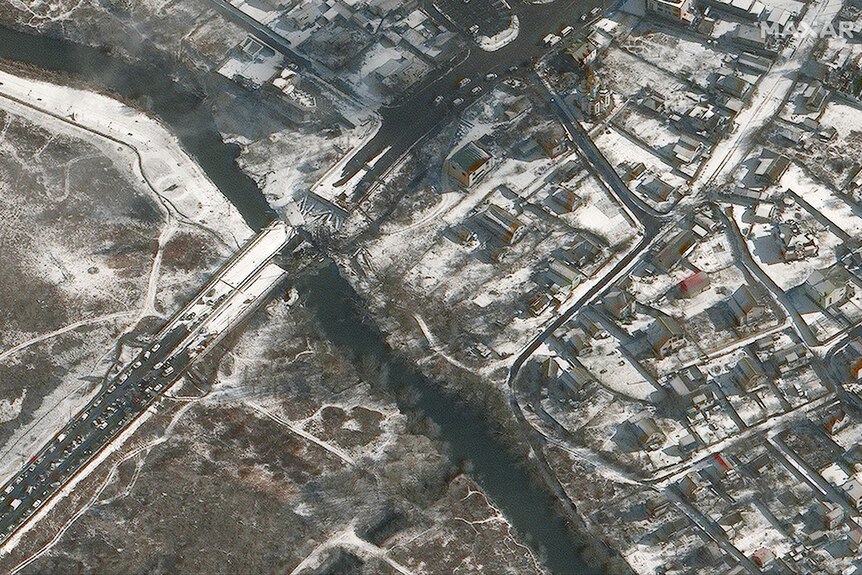 Satellite image shows destroyed bridge which ran across a river. The bridge's remains are covered in snow and waiting vehicles