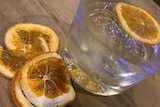 A glass of clear liquid with ice cubes and a slice of lemon on top, with more lemon slices beside the glass.