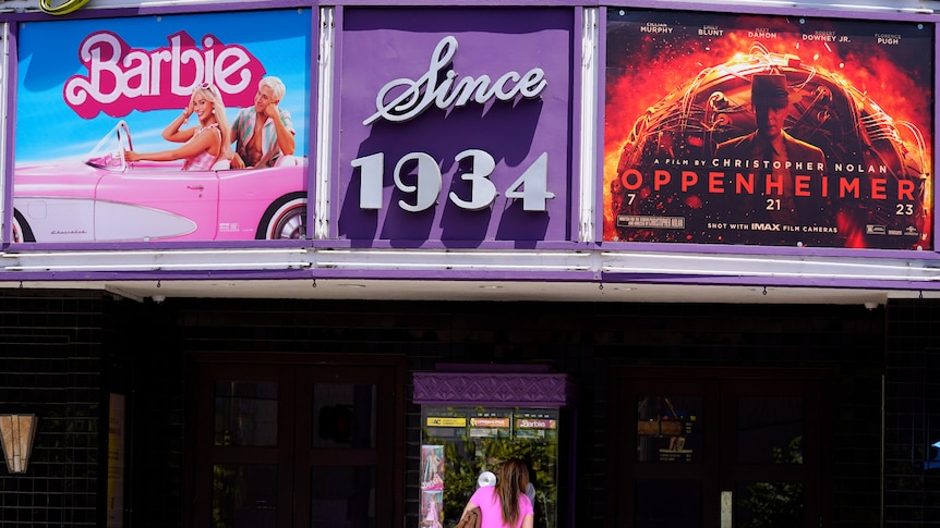 A patron buys a movie ticket underneath a marquee featuring the films "Barbie" and "Oppenheimer" at a theatre.