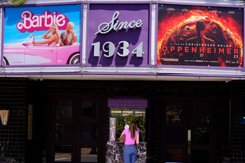 A patron buys a movie ticket underneath a marquee featuring the films "Barbie" and "Oppenheimer" at a theatre.