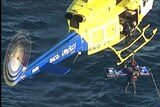 A yellow and blue helicopter lifts a body on a stretcher while flying above ocean water.