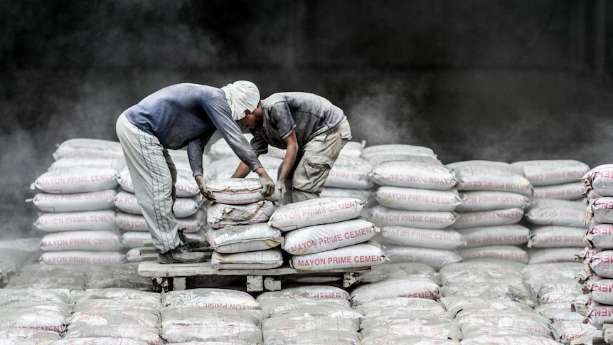 Men in blue tops hunched over pallets of cement with dust in the air.